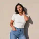 Woman with wavy dark hair in a white ribbed top and high-waisted jeans, hand resting near her neck, standing against a sunlit tan wall