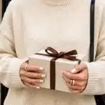 Woman in a cozy sweater holding a beautifully wrapped gift box tied with ribbon, featured in the CSPS holiday gift guide