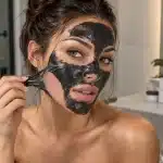 Woman peeling a black charcoal mask from her cheek in front of a bathroom mirror