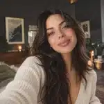 Close-up of a woman with long dark hair smiling in a cream knit cardigan at home
