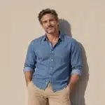 Man in a denim shirt and khakis leaning against a sunlit stucco wall